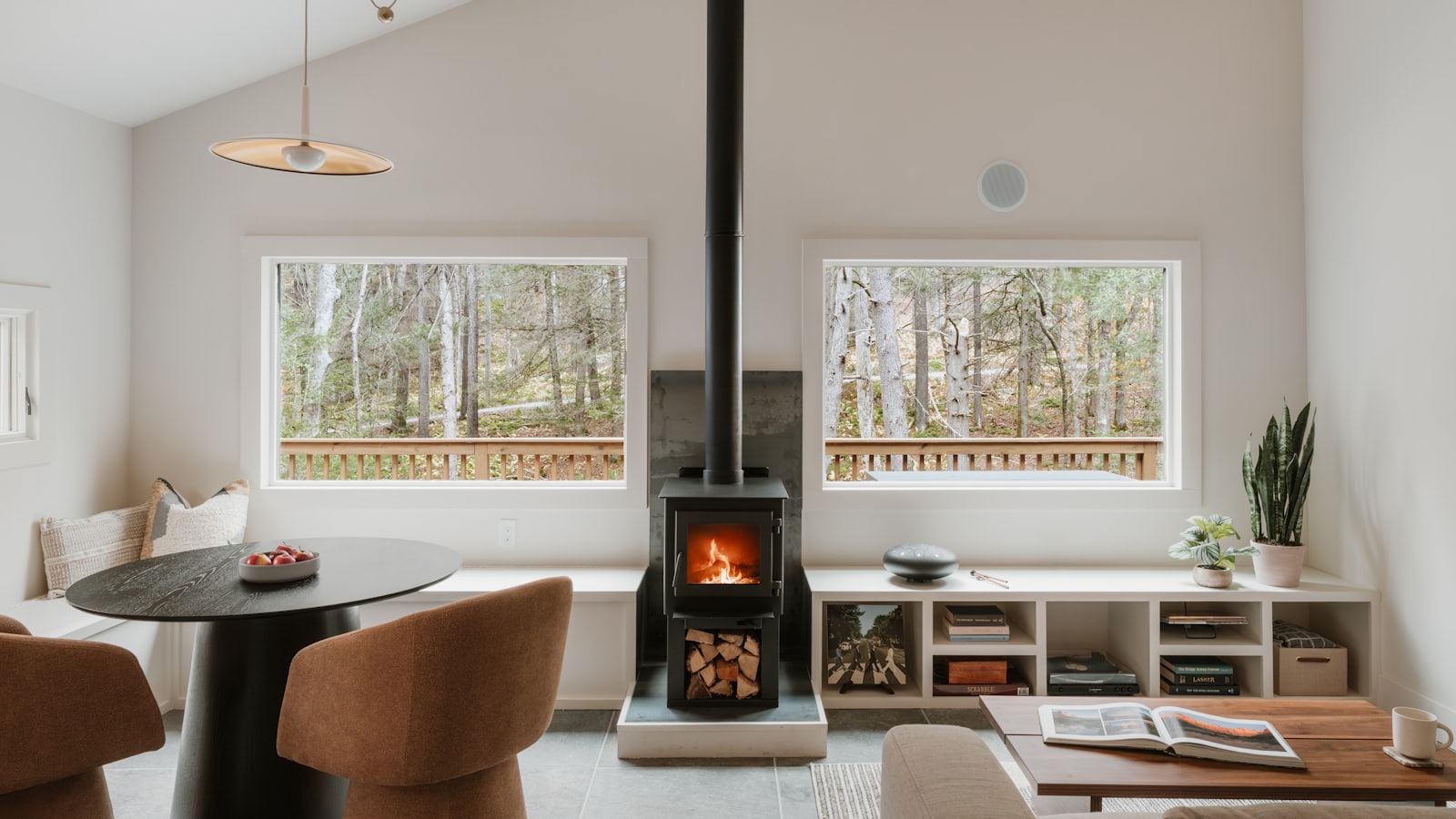 A warm cottage living room with a lit wood stove and birch woods through the windows.