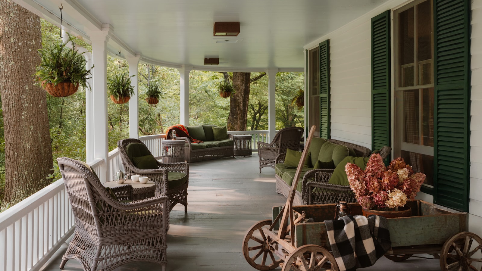 A wide covered porch with wicker chairs, hanging ferns and woods beyond.