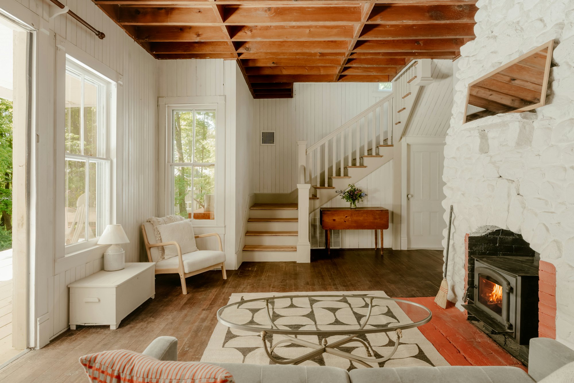 Morning light across a bright cottage living room with white board walls, wood beams and a swept hearth.